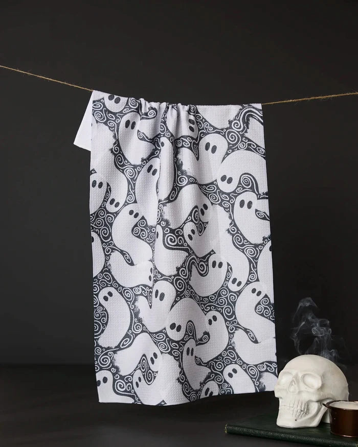 The Geometry Swirling Ghosts tea towel, featuring a cartoon ghost pattern, hangs against a dark background with a white skull and stacked books nearby for a spooky Halloween vibe.