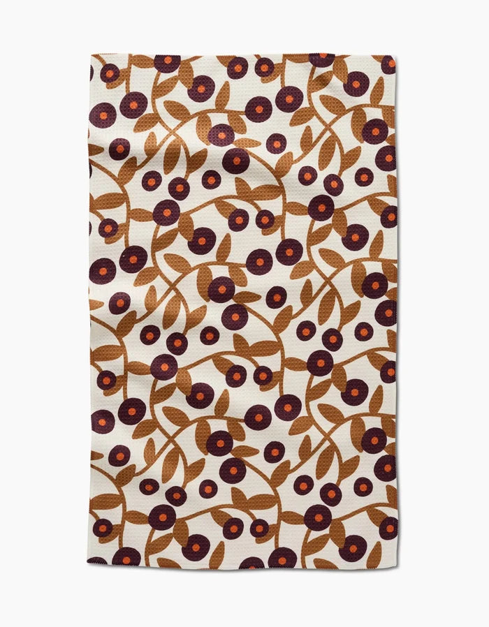The Lizzie Clark Swirling Vines tea towel is rectangular, made from recycled materials, and features brown branches with leaves and purple and red circular flowers on a cream background.
