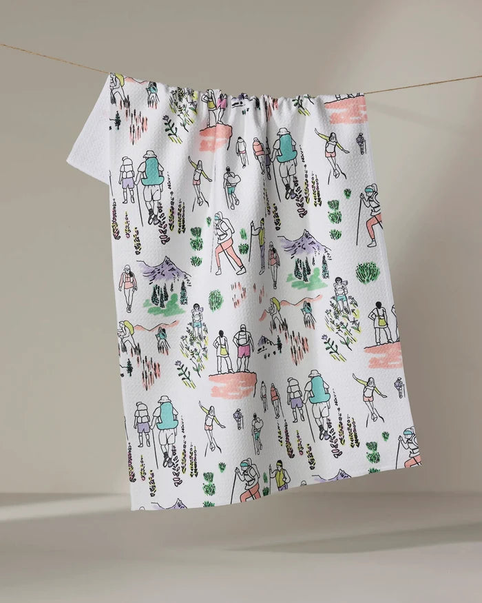 The "Take A Hike" tea towel by Elisabeth Wing features colorful, hand-drawn style illustrations of hikers, mountains, trees, and nature scenes in green, orange, purple, and yellow on white.