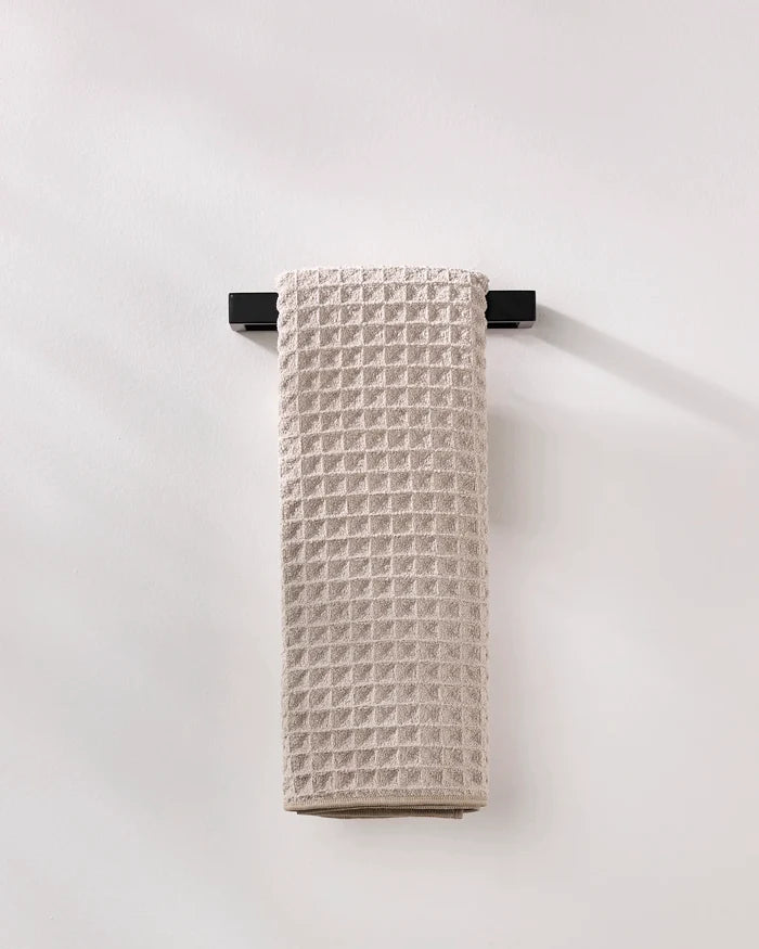 A GEOMETRY Taupe waffle hand towel made from recycled materials hangs neatly on a sleek black towel bar against a plain white wall.