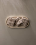 A Taupe oval plate by GEOMETRY displays three square, textured crackers with a waffle pattern—arranged side by side—on a neutral surface.
