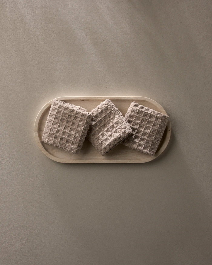 A Taupe oval plate by GEOMETRY displays three square, textured crackers with a waffle pattern—arranged side by side—on a neutral surface.