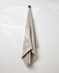 A taupe, eco-friendly GEOMETRY waffle bath towel with a textured pattern hangs neatly on a single black hook against a smooth white wall.