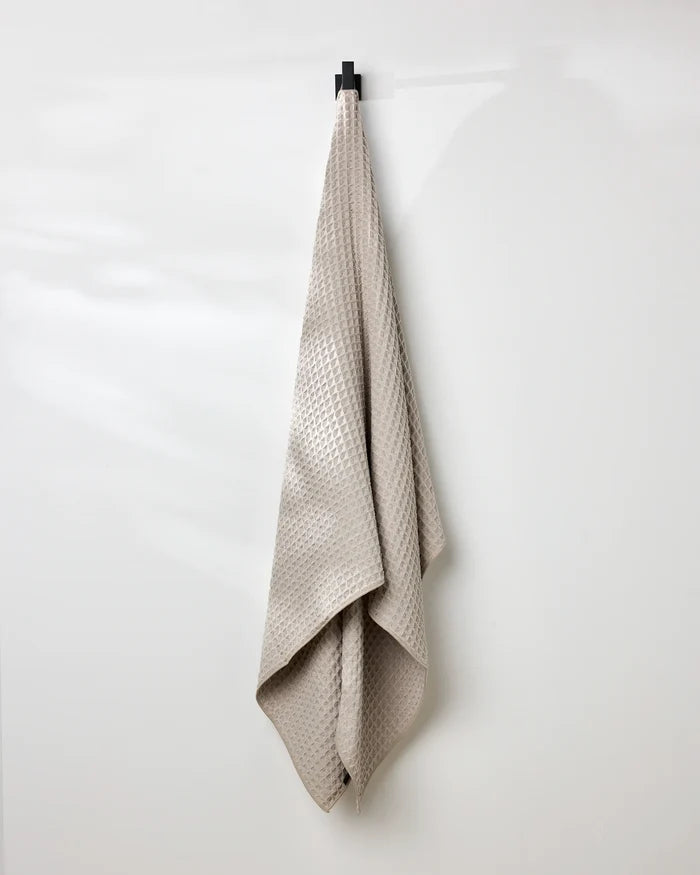 A taupe, eco-friendly GEOMETRY waffle bath towel with a textured pattern hangs neatly on a single black hook against a smooth white wall.