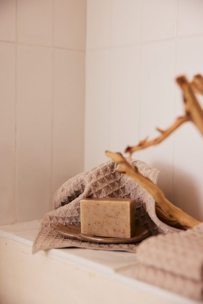 A GEOMETRY Taupe Waffle Washcloth displays a handmade bar of soap on a small dish, accented by a decorative wooden branch, all set against white tiles.