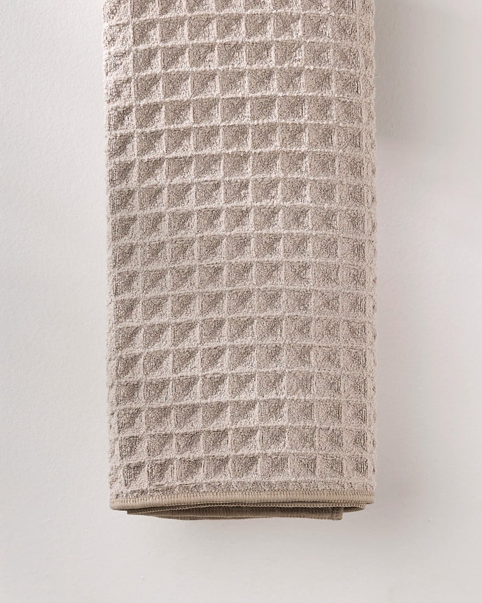 A folded GEOMETRY Taupe waffle hand towel, crafted from recycled materials, hangs against a plain white wall.
