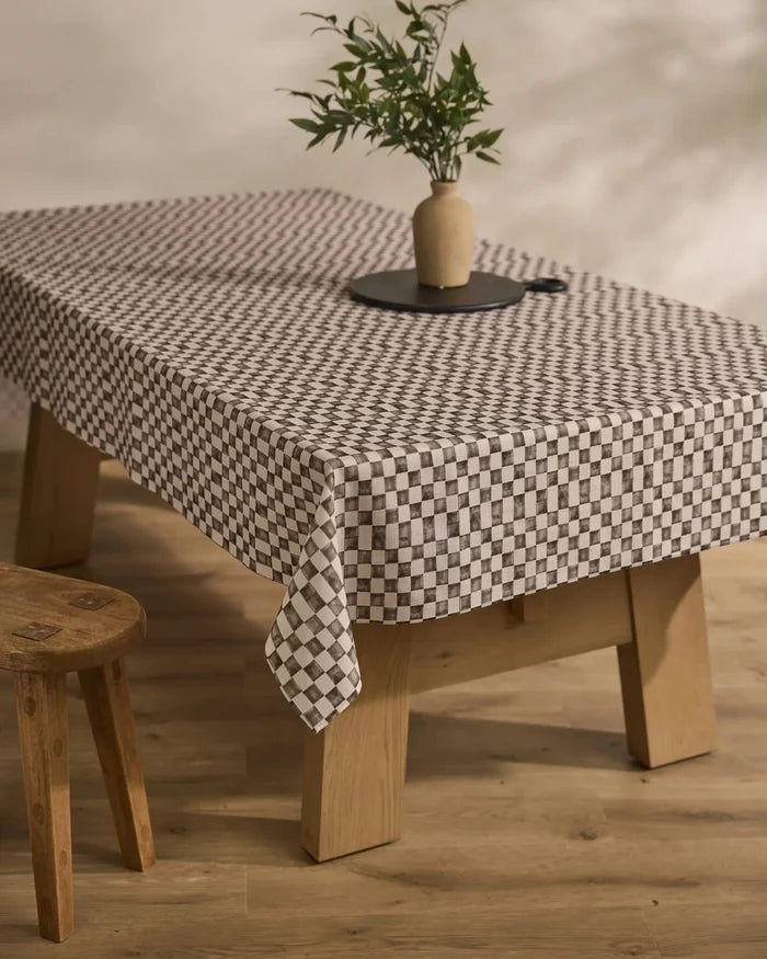 A Tavern wooden table by Geometry is topped with a stain-resistant black-and-white checkered tablecloth, a round black tray, and a ceramic vase with green leafy branches; a wooden stool sits beside it on a light wood floor.