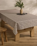 A Tavern wooden table by Geometry is topped with a stain-resistant black-and-white checkered tablecloth, a round black tray, and a ceramic vase with green leafy branches; a wooden stool sits beside it on a light wood floor.