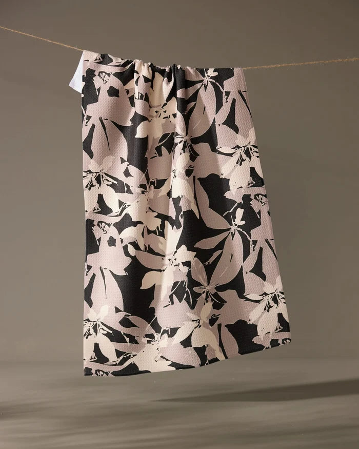 The Tawny tea towel by Geometry, featuring a beige and black abstract floral design, hangs over a thin rope against a neutral background.