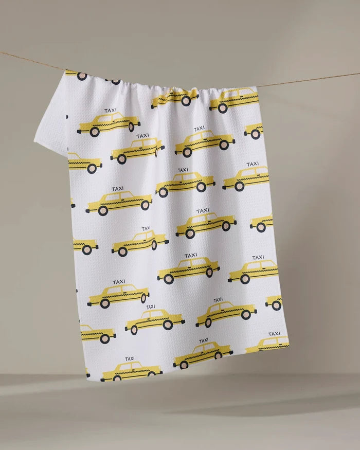 The "Taxi Cabs" tea towel by Vaishali Design Studio features a yellow taxi print and hangs on a clothesline against a neutral background.