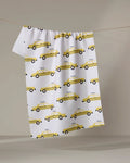 The "Taxi Cabs" tea towel by Vaishali Design Studio features a yellow taxi print and hangs on a clothesline against a neutral background.