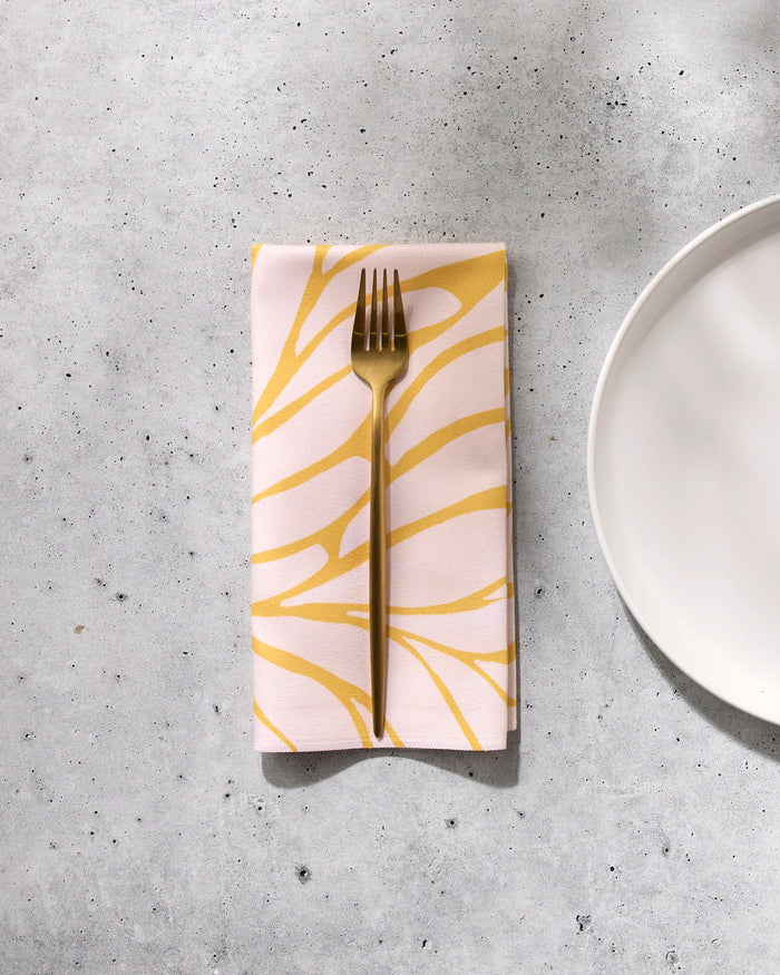 A gold fork rests on a folded GEOMETRY Tenderhearted reusable dinner napkin with a yellow abstract pattern, placed on a light gray textured surface next to a white ceramic plate.