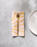 A gold fork rests on a folded GEOMETRY Tenderhearted reusable dinner napkin with a yellow abstract pattern, placed on a light gray textured surface next to a white ceramic plate.