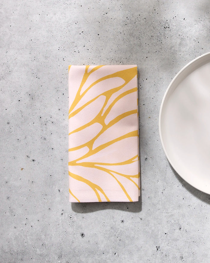 The GEOMETRY "Tenderhearted" napkin features a yellow abstract leaf pattern on a light background, folded and placed on a gray speckled surface beside the edge of a white plate.