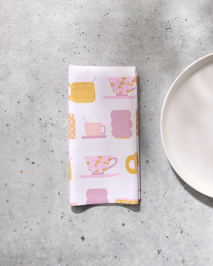 A folded white dinner napkin from the GEOMETRY Tenderhearted eco-friendly set, featuring pink and yellow teacup patterns, rests on a light gray textured surface beside a plain white plate.