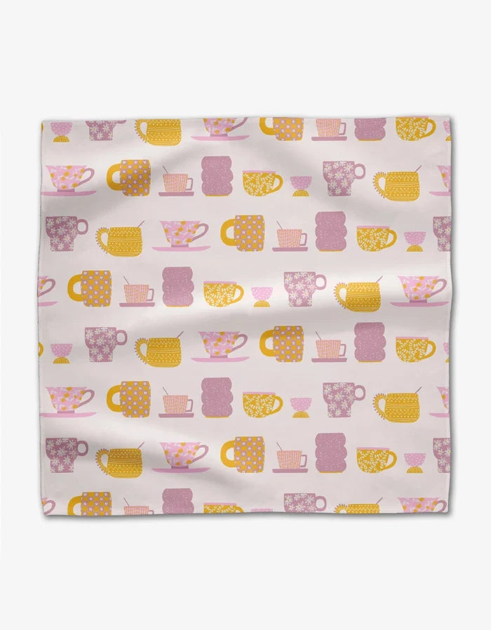 The GEOMETRY Tenderhearted is a square dinner napkin made from Geosuede fabric, showcasing a grid pattern of pink, yellow, and orange teacups, mugs, and teapots on a light background with a slightly wrinkled look.