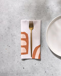 A gold fork rests on GEOMETRY’s Terracotta Vibes reusable dinner napkin, featuring abstract brown and peach patterns, folded on a light gray speckled surface beside a plain white plate.