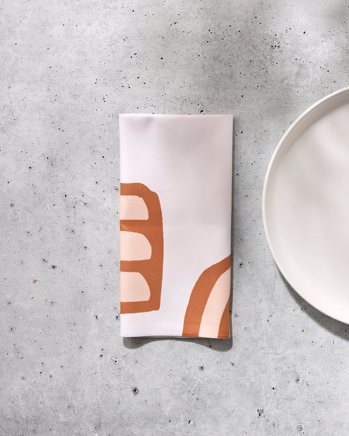 The GEOMETRY "Terracotta Vibes" folded white reusable dinner napkin with abstract orange and beige patterns rests on a speckled gray surface beside the edge of a round white plate.