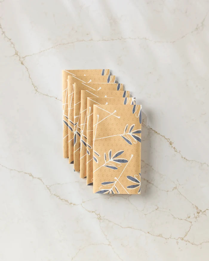 Five GEOMETRY Tess napkins, featuring a blue and white leaf pattern on tan fabric, are neatly folded and stacked on a white marble surface with light brown veining—perfect as eco-friendly kitchen towels.
