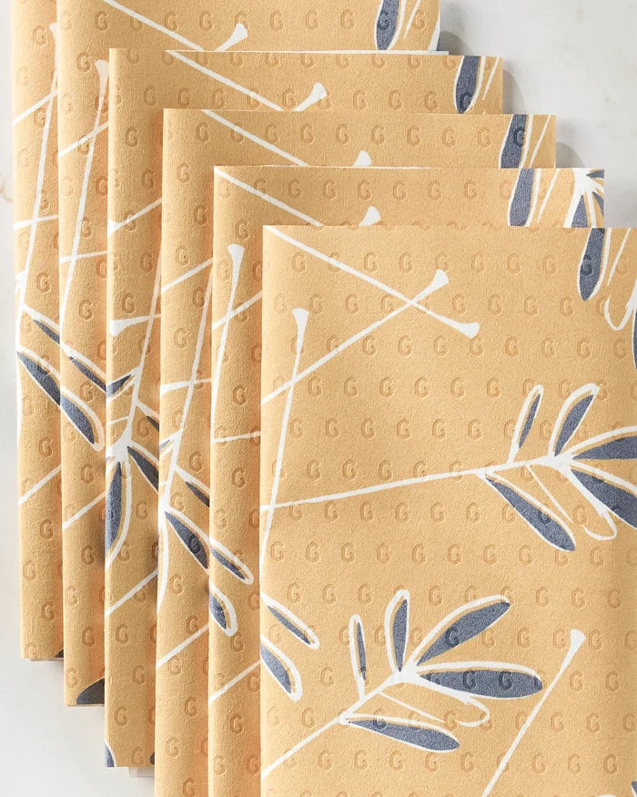 Five stacked Tess tan napkins by GEOMETRY feature a white and dark blue leaf pattern with small raised “G” letters, making them perfect eco-friendly kitchen towels for stylish, sustainable dining.