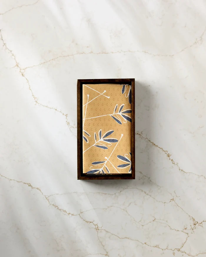 The GEOMETRY Tess rectangular soap bar, featuring a tan and blue leaf pattern, rests in a dark wooden tray on white marble beside eco-friendly kitchen towels, enhancing the elegant atmosphere.
