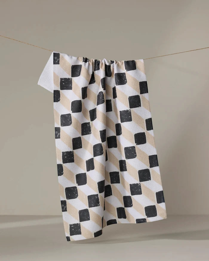 The Tessera kitchen towel by Studio Jeannot, crafted from recycled materials, features a geometric black, beige, and off-white cube pattern and hangs on a string against a plain light background.