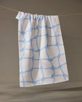 The Geometry Tethered Blue ultra-absorbent dish towel, featuring a blue rope knot pattern on white, hangs from a beige string against a neutral background.