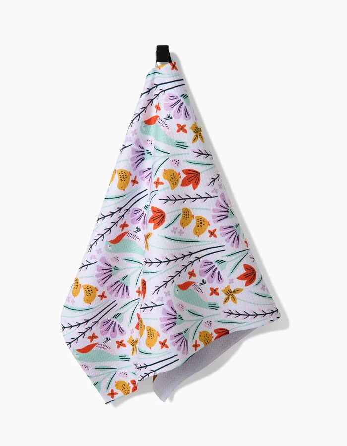 The Thaddeus kitchen towel by Amy MacCready features a white base with orange, yellow, green, and purple floral patterns. Made from recycled materials, it hangs from a black loop against a plain white background.
