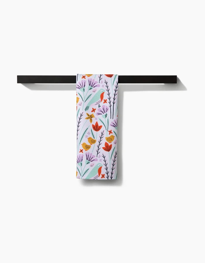 The Amy MacCready “Thaddeus” tea towel, featuring a vibrant floral and bird design, hangs neatly on a black horizontal towel bar against a white background.