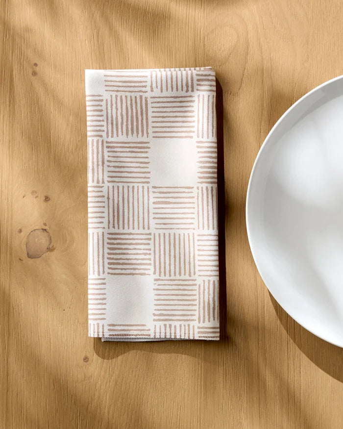 The Thatcher napkin by Studio Jeannot, featuring a beige grid pattern and eco-friendly recycled materials, rests folded on a light wooden table beside a white ceramic plate, sunlight softly highlighting its elegant charm.