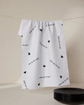 A white ultra-absorbent tea towel by GEOMETRY, called The Best, featuring small black hearts and MOMS ARE THE BEST print, hangs above black plates.