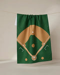 The Field tea towel by GEOMETRY, made from recycled materials, displays a brown and white baseball diamond illustration on green fabric and hangs against a neutral background.