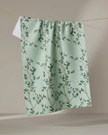 The Lily by Becki Owens is a light green kitchen tea towel featuring a branch and leaf pattern with pink flowers. Made from recycled materials, it’s an eco-friendly and charming accent for your home.