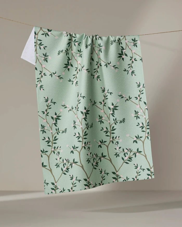 The Lily by Becki Owens is a light green kitchen tea towel featuring a branch and leaf pattern with pink flowers. Made from recycled materials, it’s an eco-friendly and charming accent for your home.