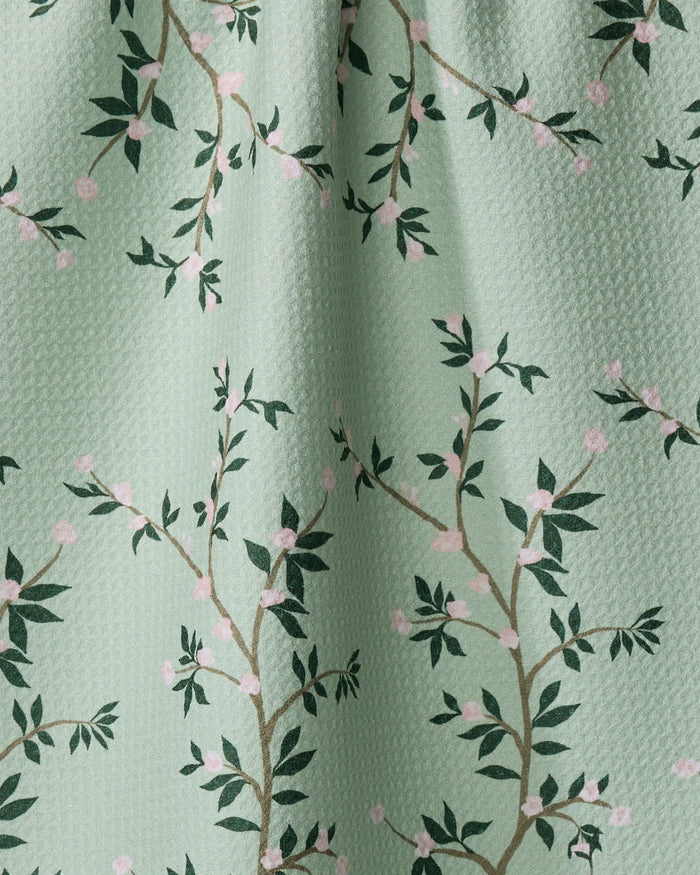 A close-up of The Lily by Becki Owens shows light green textured fabric with dark green leafy branches and pink flowers. Gentle folds highlight its ultra-absorbent quality, making it perfect as a kitchen tea towel.