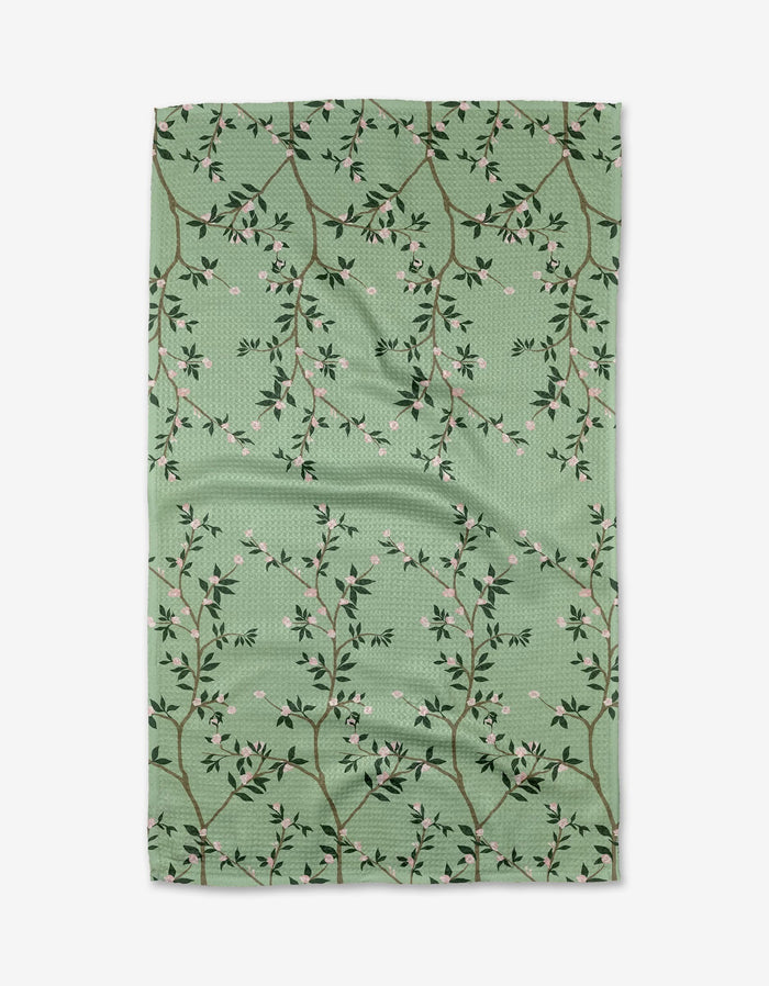 The Lily Tea Towel by Becki Owens features a green Geoweave™ fabric with branches, small leaves, and tiny pink flowers in a lattice pattern, shown slightly wrinkled against a white background.