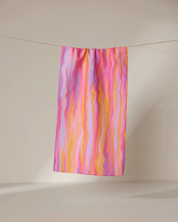 The Rush quick-drying towel by GEOMETRY, featuring wavy pink, yellow, and purple stripes, hangs from a clothesline against a plain beige background.