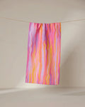 The Rush quick-drying towel by GEOMETRY, featuring wavy pink, yellow, and purple stripes, hangs from a clothesline against a plain beige background.