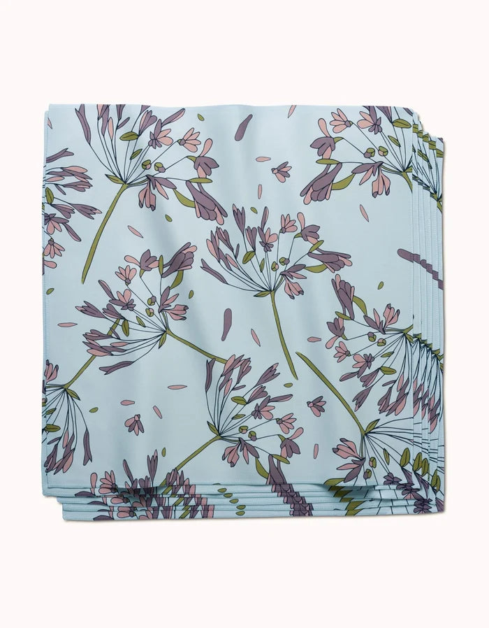 The Wind by GEOMETRY: a folded, light blue reusable dinner napkin with a botanical print of delicate purple, pink, and green flowers and leaves.