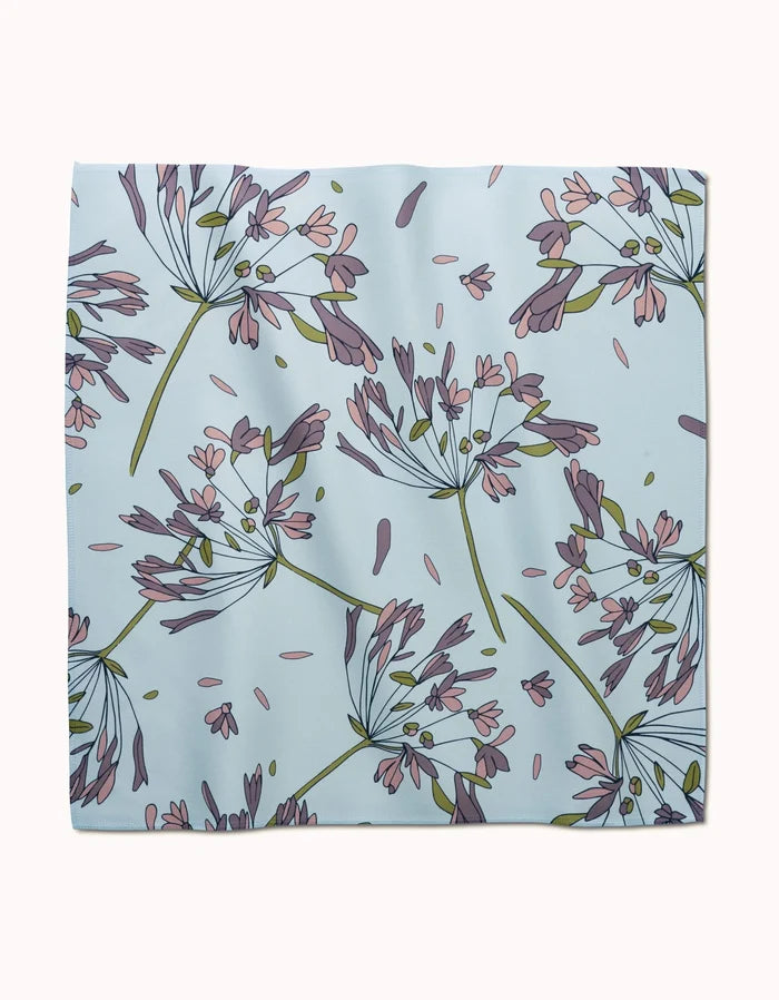 The Wind by GEOMETRY is a square, light blue reusable dinner napkin with a floral design of purple and pink flowers with green stems and leaves in various directions.
