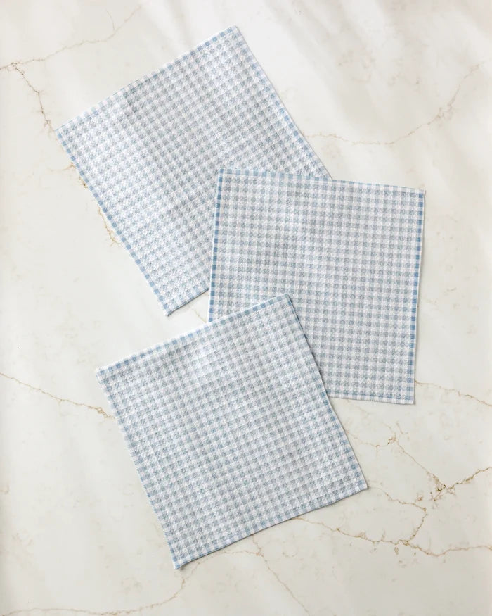 Three Tidepool Check dishcloths by Jenny Keller, featuring a light blue and white houndstooth pattern, are neatly displayed on a white marble surface with subtle beige veining.
