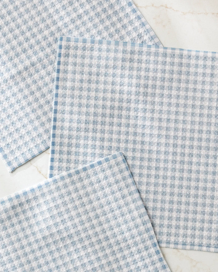 Three Tidepool Check cloths by Jenny Keller, featuring a light blue and white checkered textured pattern and made from recycled materials, are laid out overlapping at the corners on a white marble surface.
