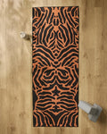 The GEOMETRY Tiger yoga mat with an orange and black pattern is on a wooden floor. At the top left is a beige water bottle, and at the bottom right are a slip-resistant yoga towel and light gray yoga blocks.