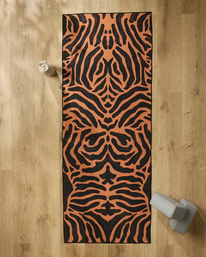 The GEOMETRY Tiger yoga mat with an orange and black pattern is on a wooden floor. At the top left is a beige water bottle, and at the bottom right are a slip-resistant yoga towel and light gray yoga blocks.