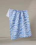 The Tinker kitchen towel by Sissy Moon, featuring a blue and white abstract wavy design, hangs on a thin clothesline against a neutral background.