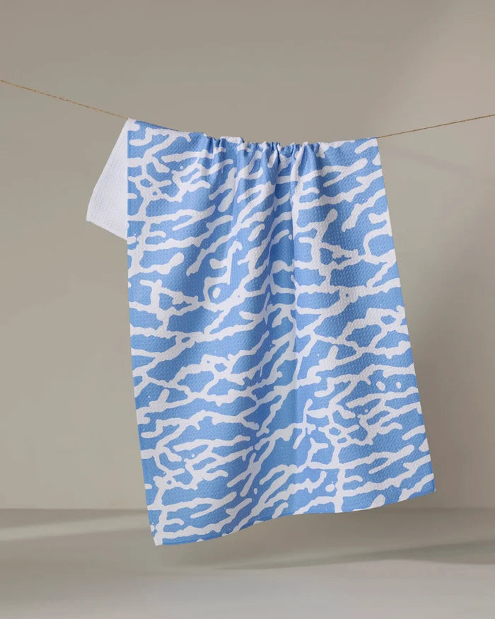 The Tinker kitchen towel by Sissy Moon, featuring a blue and white abstract wavy design, hangs on a thin clothesline against a neutral background.