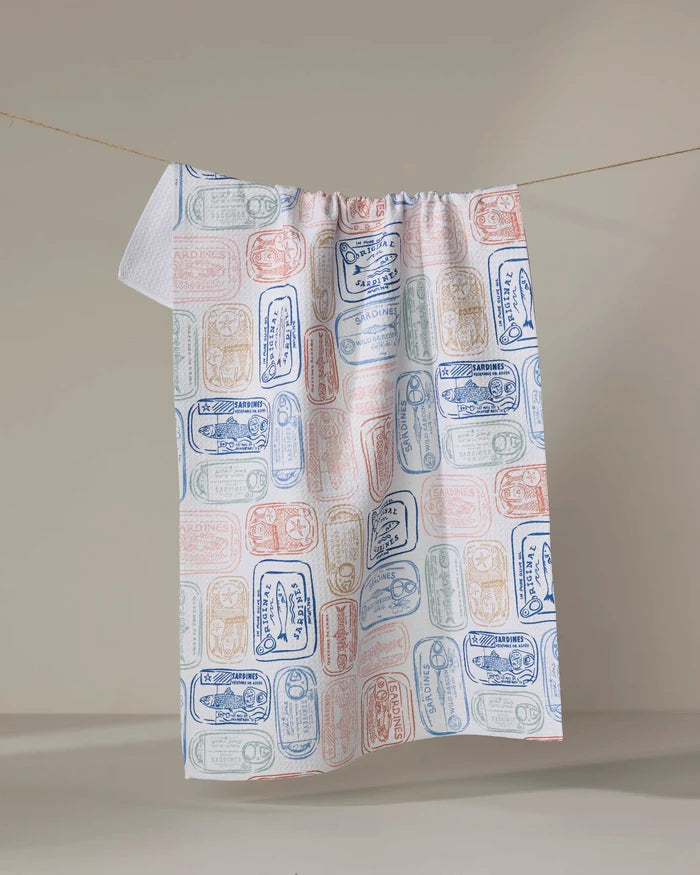 The "Tinned Sardines" dish towel by Hufton Studio features a vintage-inspired sardine can label pattern in blue, red, and green on white, shown hanging on a line against a neutral background.