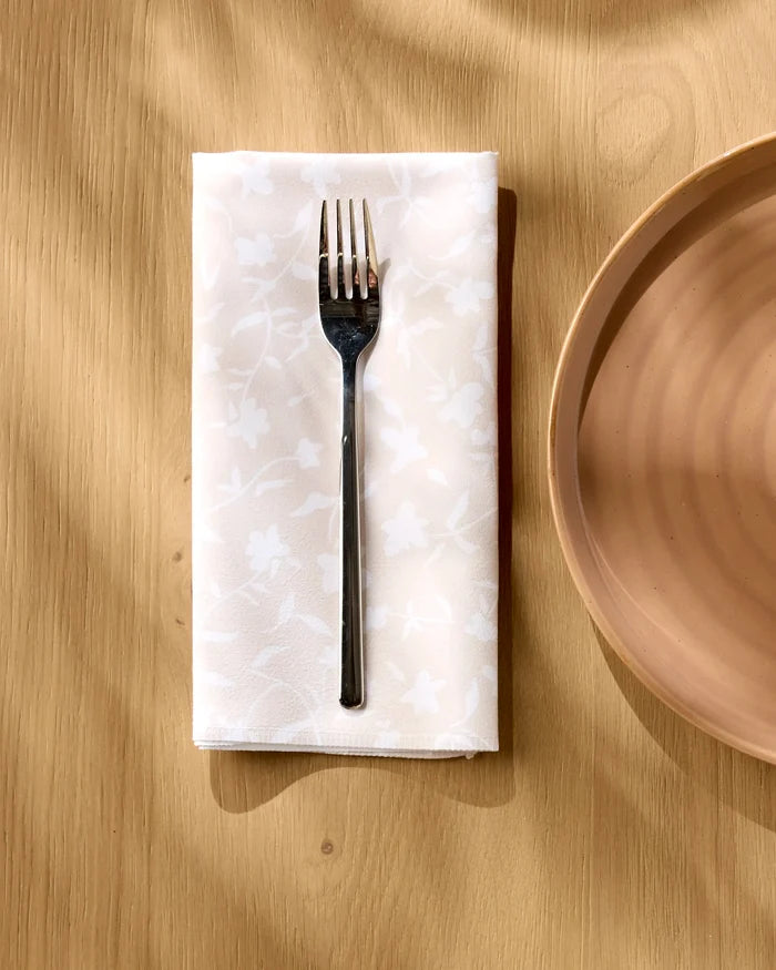 A silver fork rests on an Aubrey Fairchild "Tiny Floral" reusable napkin, folded on a light wooden table beside a round beige plate.