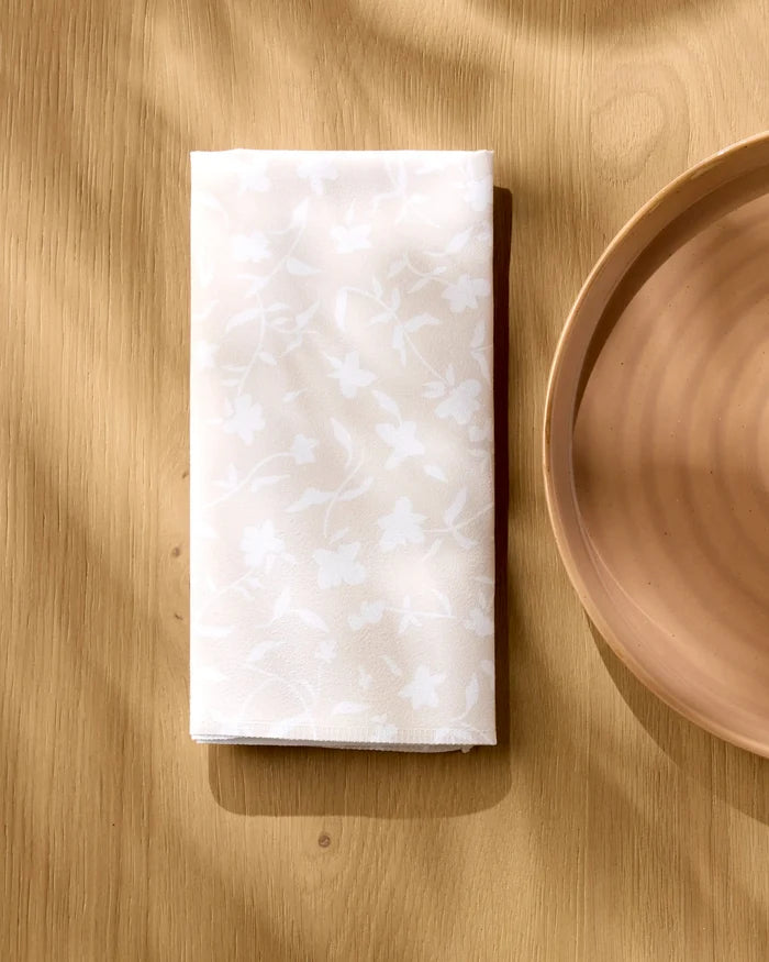 The Aubrey Fairchild Tiny Floral white reusable dinner napkin, featuring a subtle floral pattern, is folded on a light wooden table beside a round beige plate, illuminated by soft natural light and gentle shadows.
