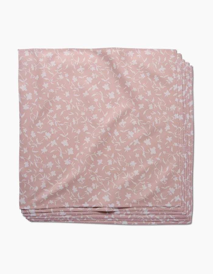 A neatly folded, pale pink Tiny Floral dinner napkin by Aubrey Fairchild, made from soft Geosuede fabric with a white floral pattern, is stacked with identical napkins on a white background.
